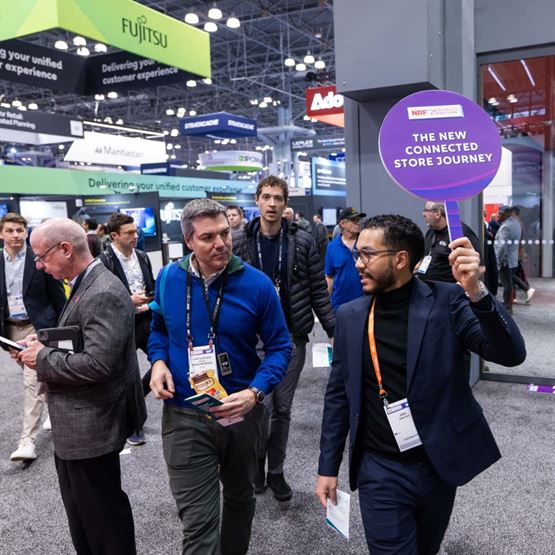 Visite guidée du salon NRF, un intervenant tient un panneau "The New Connected Store Journey" en tête de groupe, au milieu des exposants.