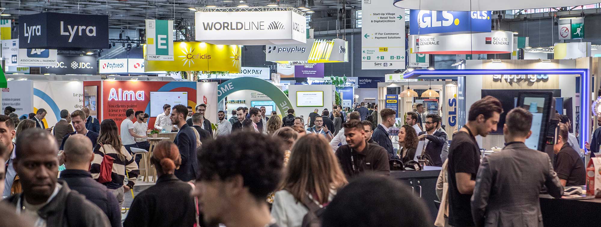 Allées animées du salon avec des visiteurs et des stands tels que Lyra, Worldline, GLS et Alma.