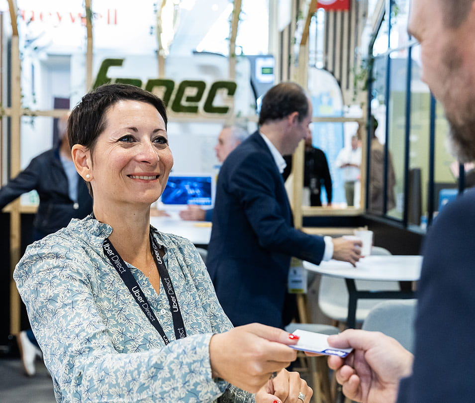 Femme souriante tendant une carte de visite lors d'une discussion sur un stand professionnel.