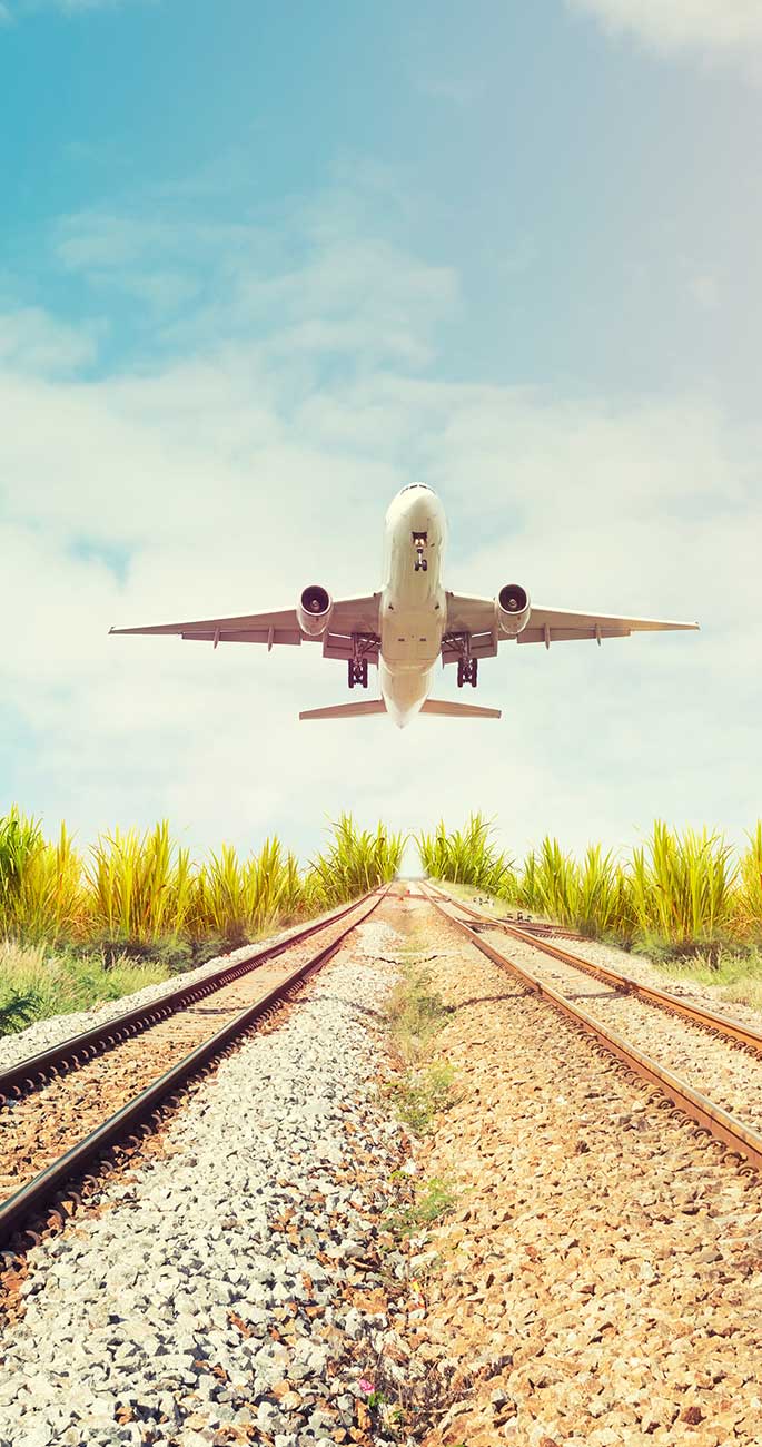 Airplane taking off above railway tracks surrounded by greenery and blue sky.