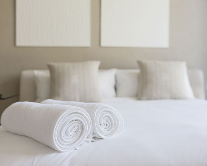 Comfortable bed in a hotel room with rolled white towels and beige cushions.