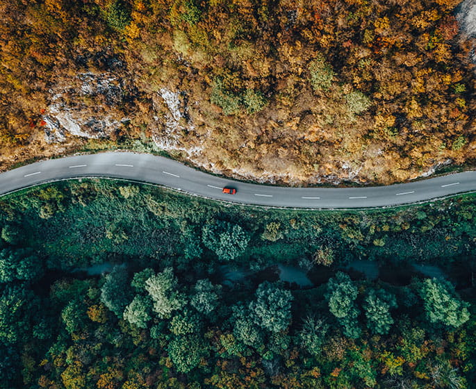 Aerial view of a winding road surrounded by trees and autumn-colored hills, with a red car in motion.